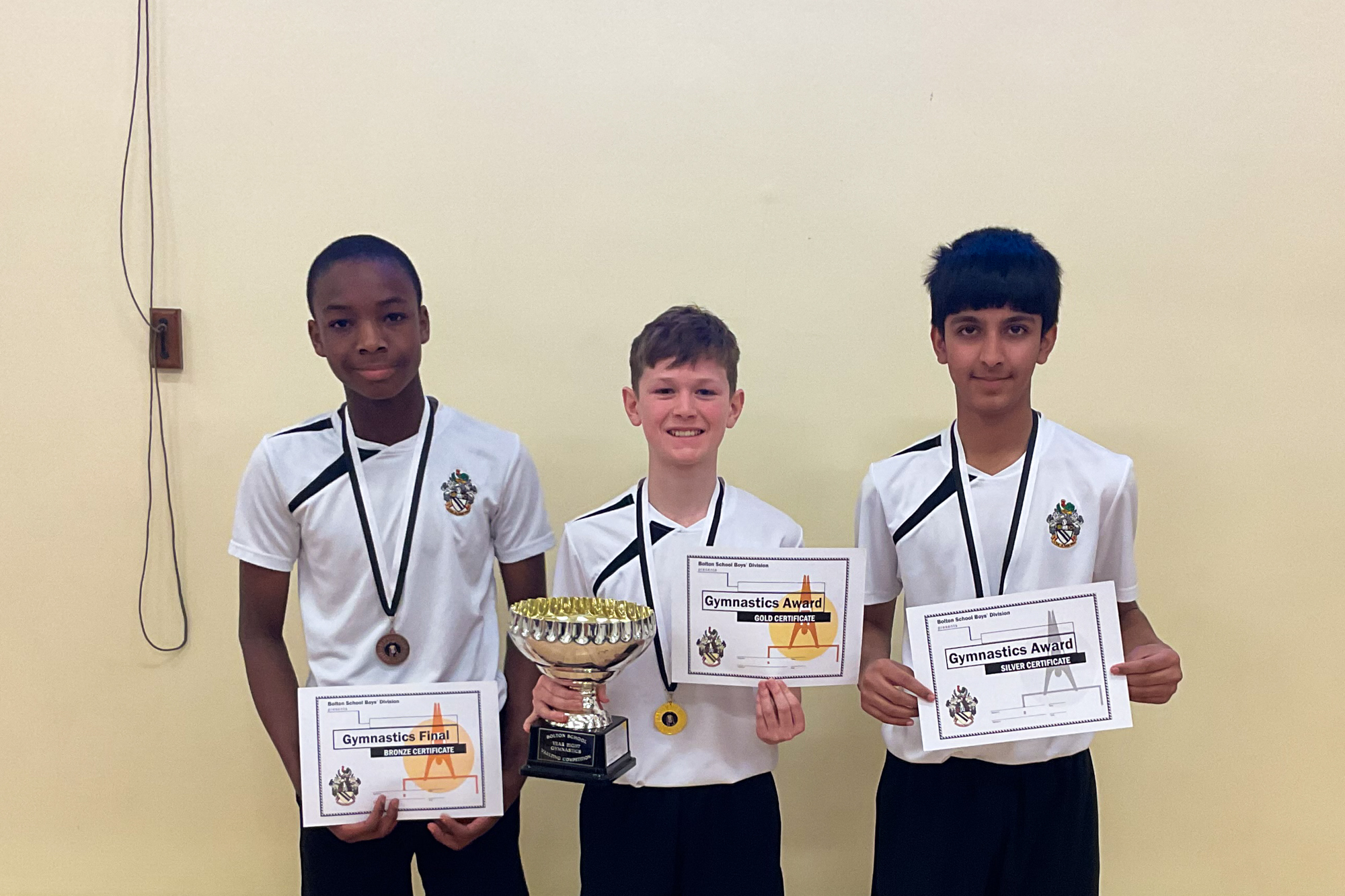 Three diverse boys in black and white PE kit, holding bronze (left) silver (right) and gold (middle) certificates and wearing matching medals. The centre boy is holding a gold cup trophy.