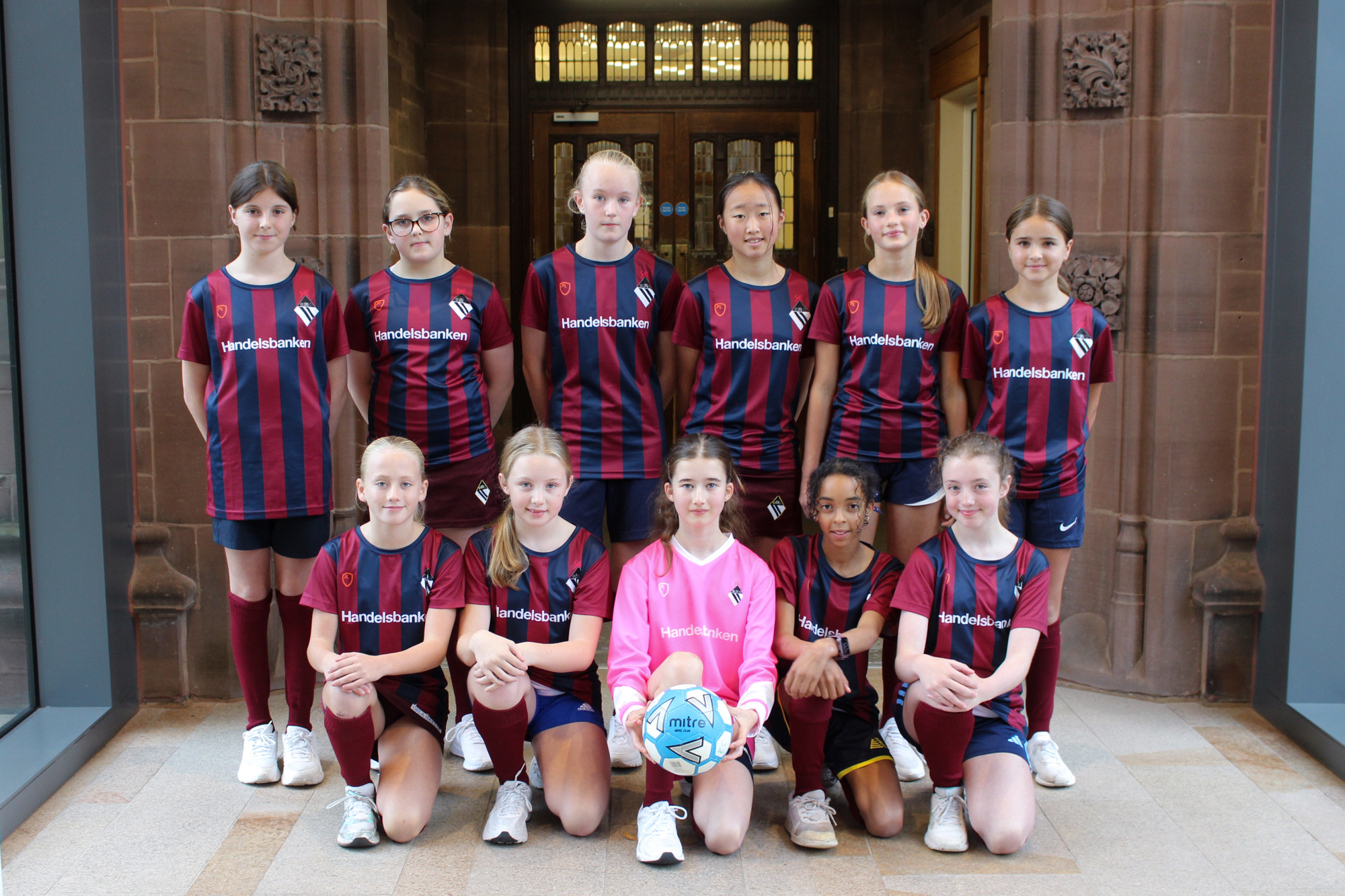 The Bolton School Girls' Division Under 13 football team: 11 girls wearing Handelsbanken sponsored Girls' Division football shirts featuring the Girls' Division crest on the chest, Handelsbanken across the chest, and vertical maroon and dark blue stripes. The goalie is wearing a solid pink shirt with the same school crest and sponsor logo. 6 girls are standing on the back row with hands behind their backs, and 5 are on one knee in front with the goalie in the centre holding a ball.