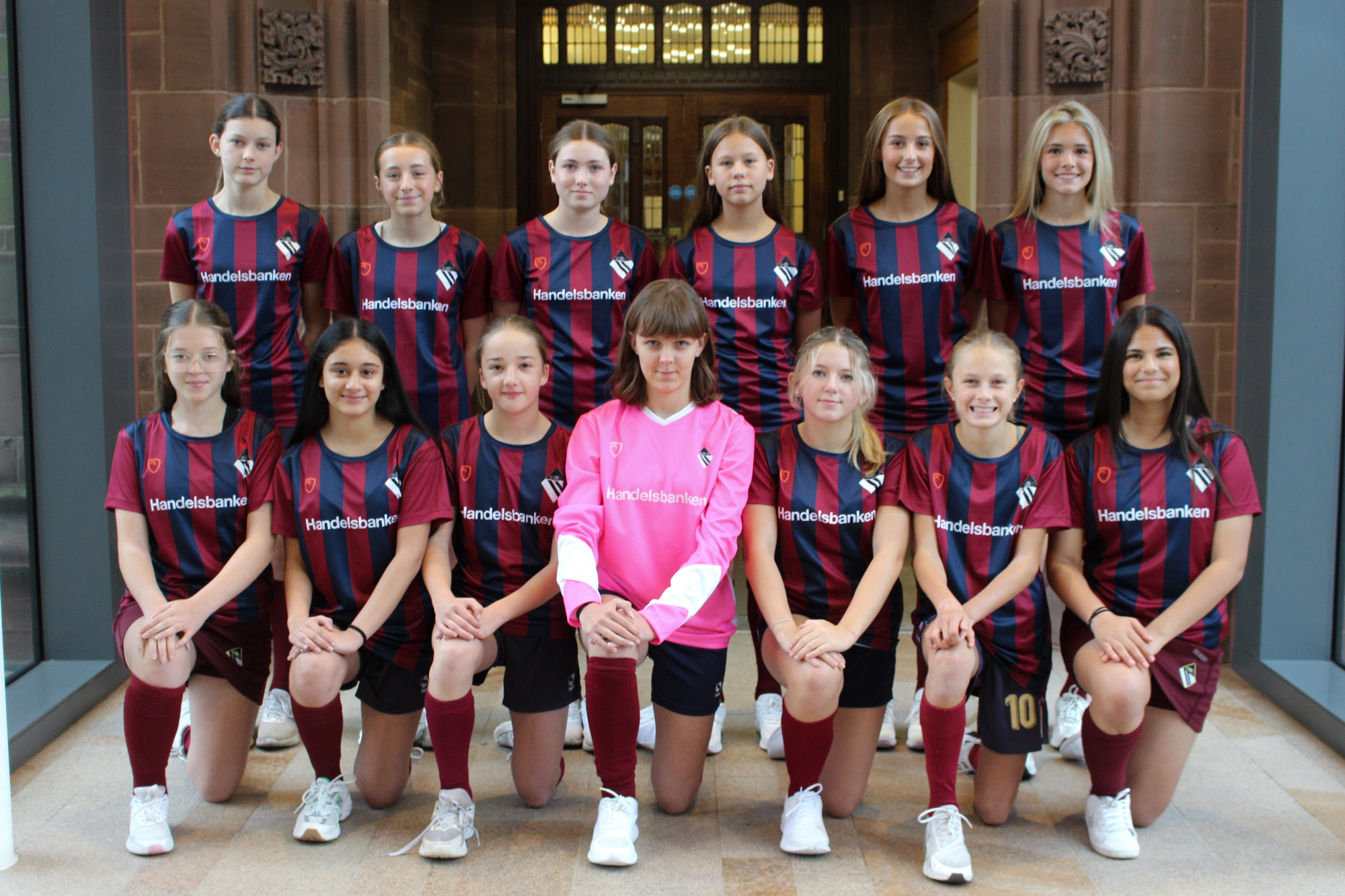 The Bolton School Girls' Division Under 15 football team: 13 girls wearing Handelsbanken sponsored Girls' Division football shirts featuring the Girls' Division crest on the chest, Handelsbanken across the chest, and vertical maroon and dark blue stripes. The goalie is wearing a solid pink shirt with the same school crest and sponsor logo. 6 girls are standing on the back row with hands behind their backs, and 7 are on one knee in front with the goalie in the centre.