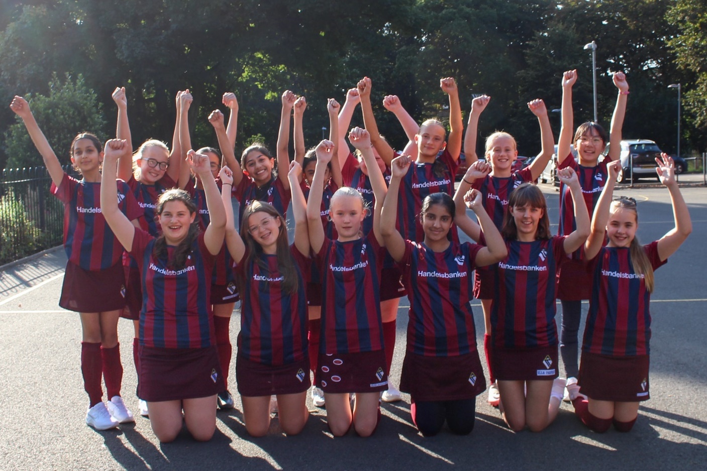 Members of the two Girls' Division Under football teams cheering while wearing Handelsbanken sponsored Girls' Division football shirts: the Girls' Division crest is on the chest, Handelsbanken is written across the chest, and the pattern is vertical maroon and dark blue stripes