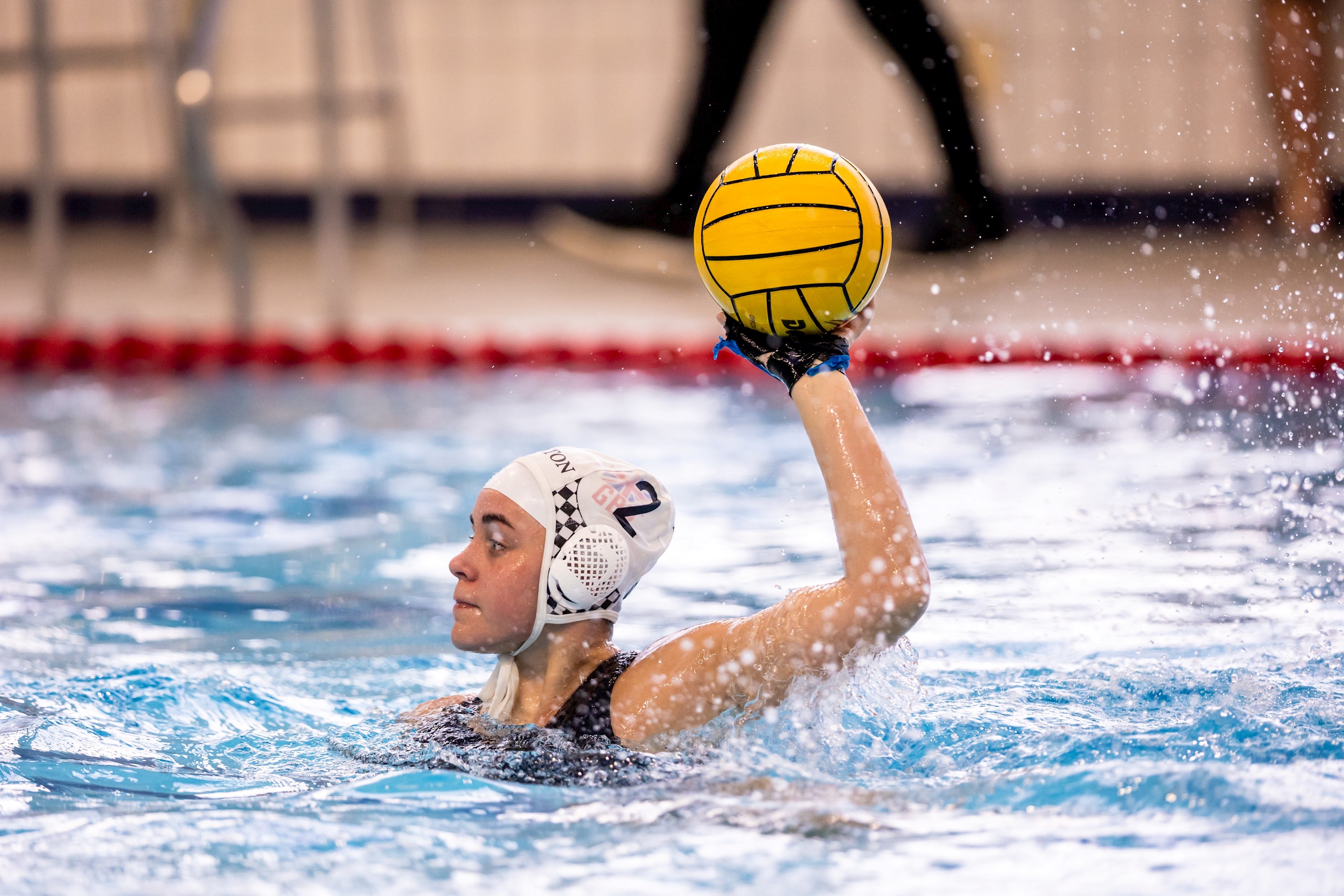 A water polo player with a 2 on the side of her white cap, submerged in the pool to her shoulders, with her left arm raised out of the water ready to throw the bright yellow ball held in her hand