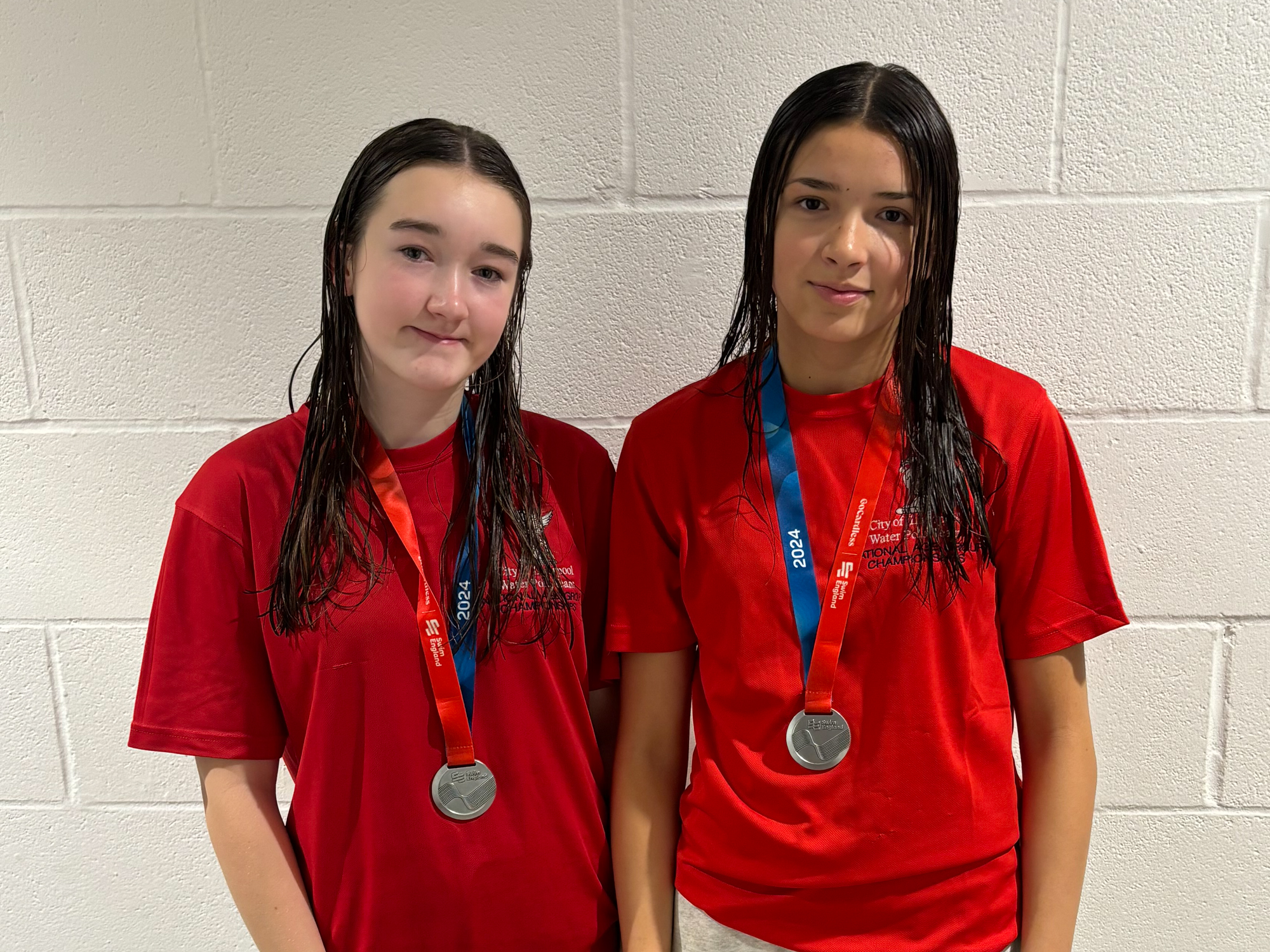 Two girls with wet hair standing shoulder to shoulder, wearing silver medals over their red City of Liverpool Water Polo National Age Group Championships polo shirts