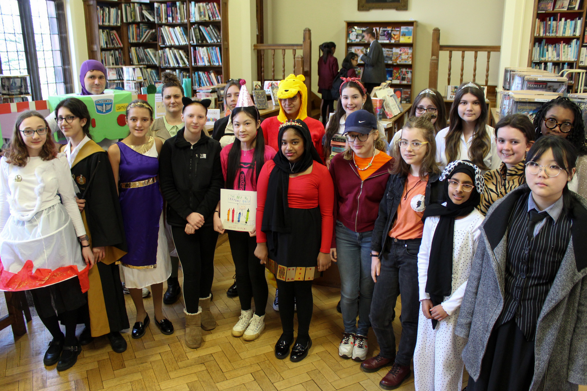 A large group of Girls' Division pupils dressed in colourful book character costumes amidst the shelves in the large, spacious Library