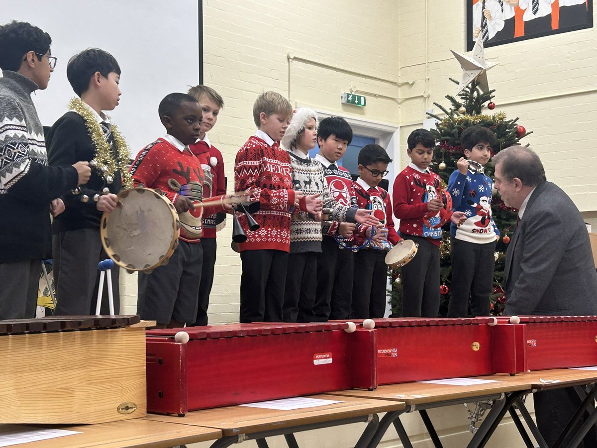 A diverse group of Bolton School Junior Boys' School pupils wearing brightly coloured Christmas jumpers and holding percussion instruments (including tambourines, agogo bells and jingle bells) stand along the front of a stage beside a Christmas Tree while performing