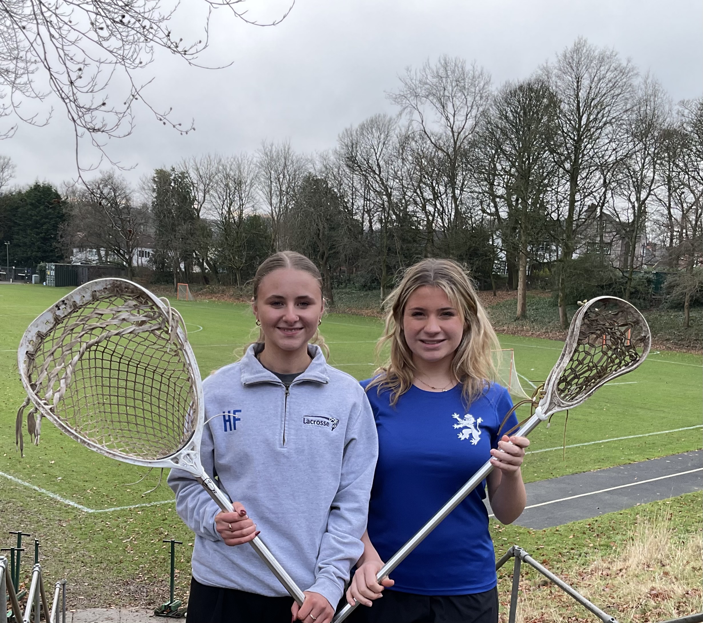 Two Bolton School girls, one holding a lacrosse goalie's stick and the other a lacrosse stick, wearing Scotland kit in front of the school field