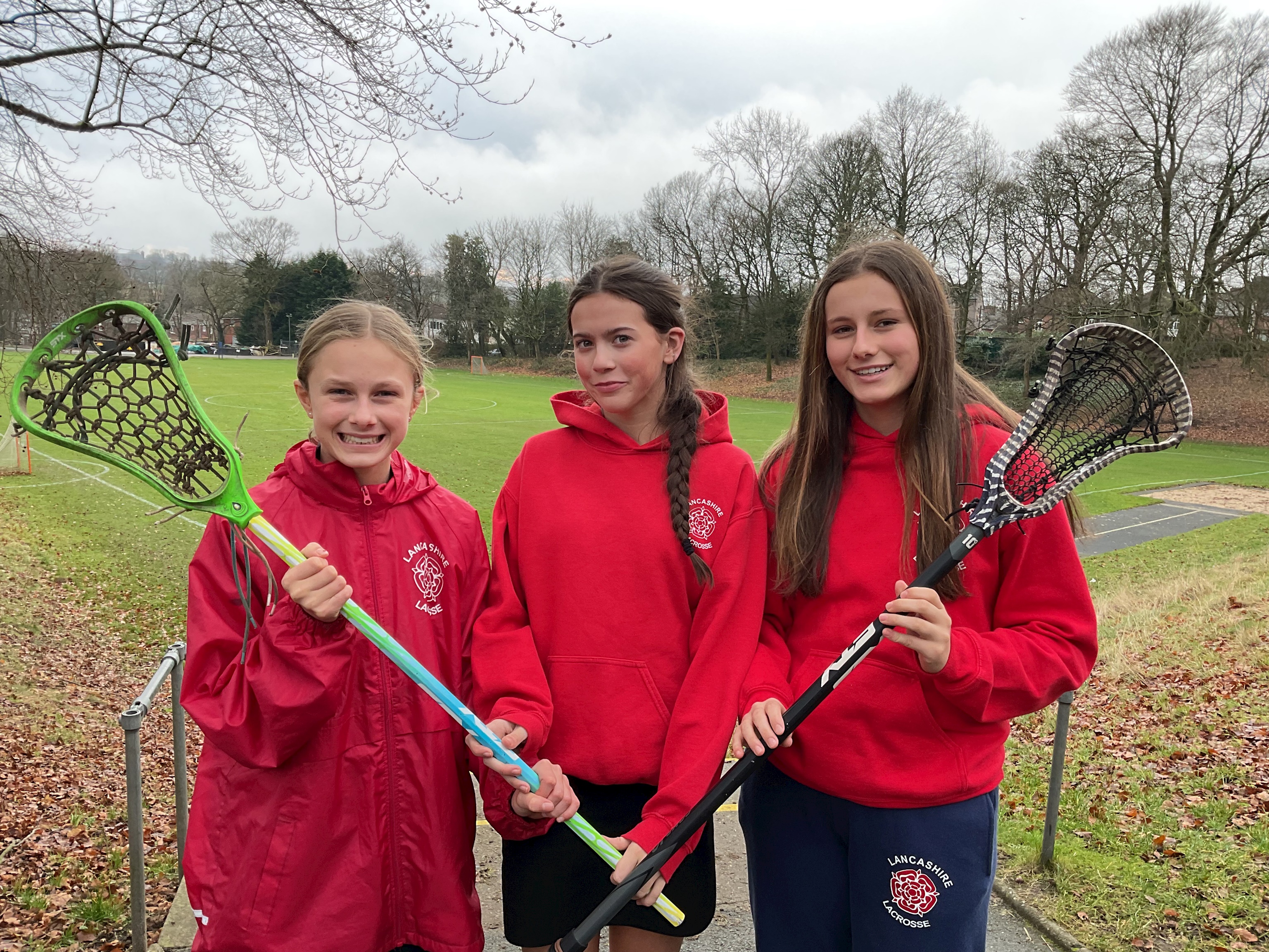 Three Bolton School girls wearing red 'Lancashire Lacrosse' hoodies and holding Lacrosse sticks and standing in front of the school field