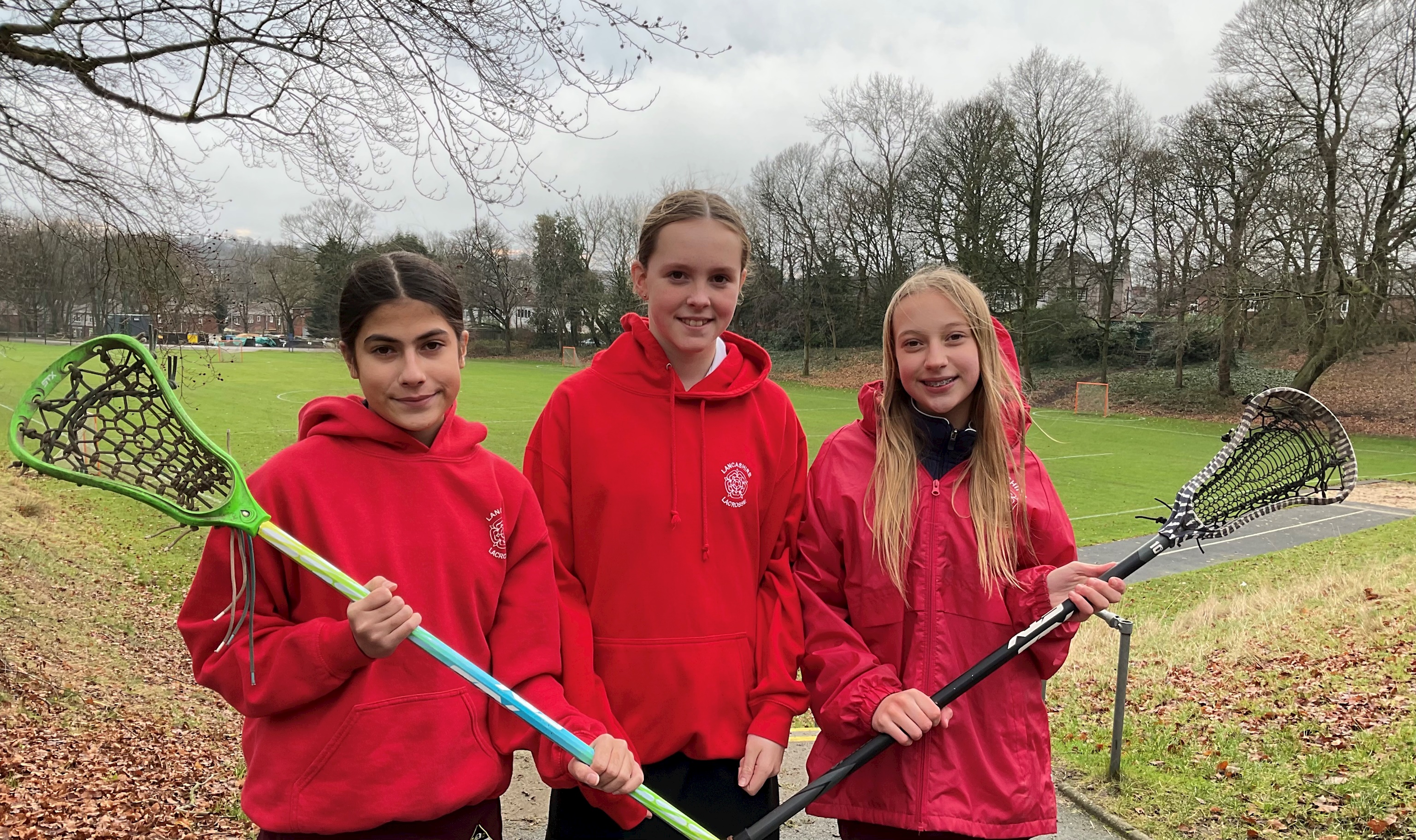 Three Bolton School girls wearing red 'Lancashire Lacrosse' hoodies and holding Lacrosse sticks and standing in front of the school field